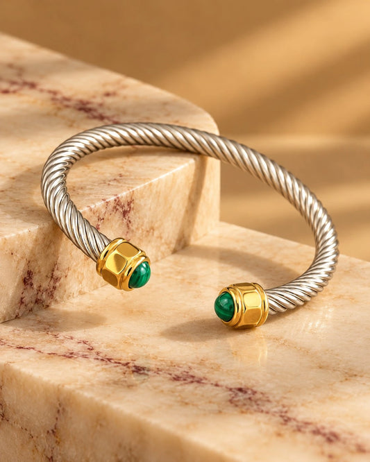 The Malachite Cable Torque Cuff Bangle resting on stacked travertine marble blocks - angled view showing twisted rope cable texture and both malachite-tipped gold ends in warm afternoon light