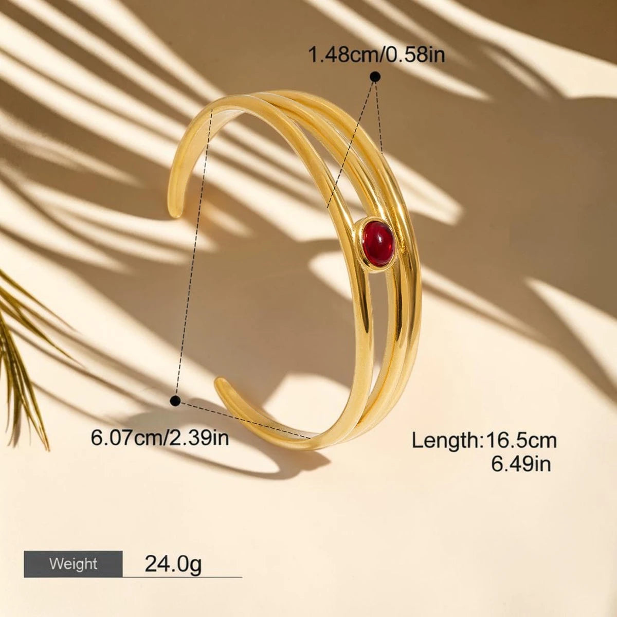 Gold-colored wrap-style bangle bracelet with a red oval stone accent, shown with dimension callouts, length, and weight text on a beige background with palm leaf shadows.