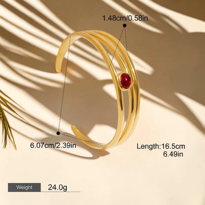 Gold-colored wrap-style bangle bracelet with a red oval stone accent, shown with dimension callouts, length, and weight text on a beige background with palm leaf shadows.