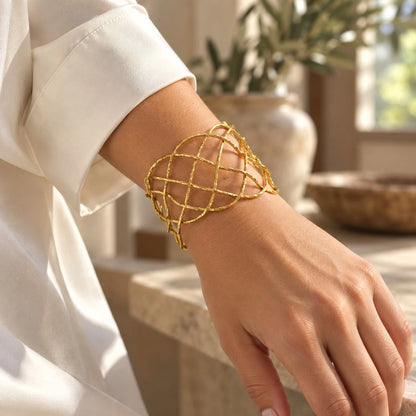 The Lattice Gold Open-Weave Wide Cuff Bangle — front product shot showing diagonal diamond-grid wire lattice structure in warm 18K gold on cream background with palm leaf shadow