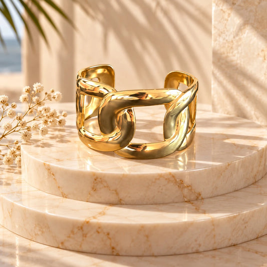 The Interlock Gold Cuban Link Wide Open Cuff Bangle worn on model in burgundy satin gown at chandelier-lit gala holding champagne coupe — close-up of wrist showing bold gold Cuban link cuff catching candlelight