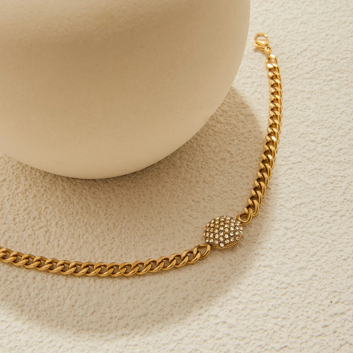 The Orbit Gold Pavé Crystal Ball Chain Bracelet — close-up flat lay on cream surface showing crystal ball charm and Cuban link chain texture