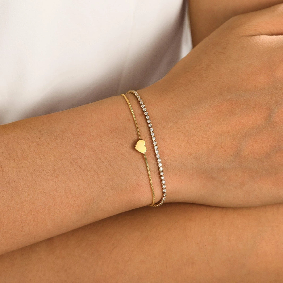 The Duo Gold Heart Charm Double Layer Bracelet — flat lay on brushed steel surface showing crystal tennis chain layered over snake chain with gold heart charm