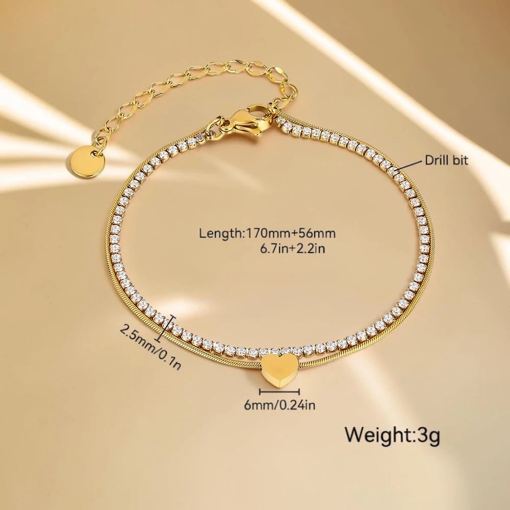 The Duo Gold Heart Charm Double Layer Bracelet — flat lay on peach surface with seashell prop showing tennis and snake chain layers