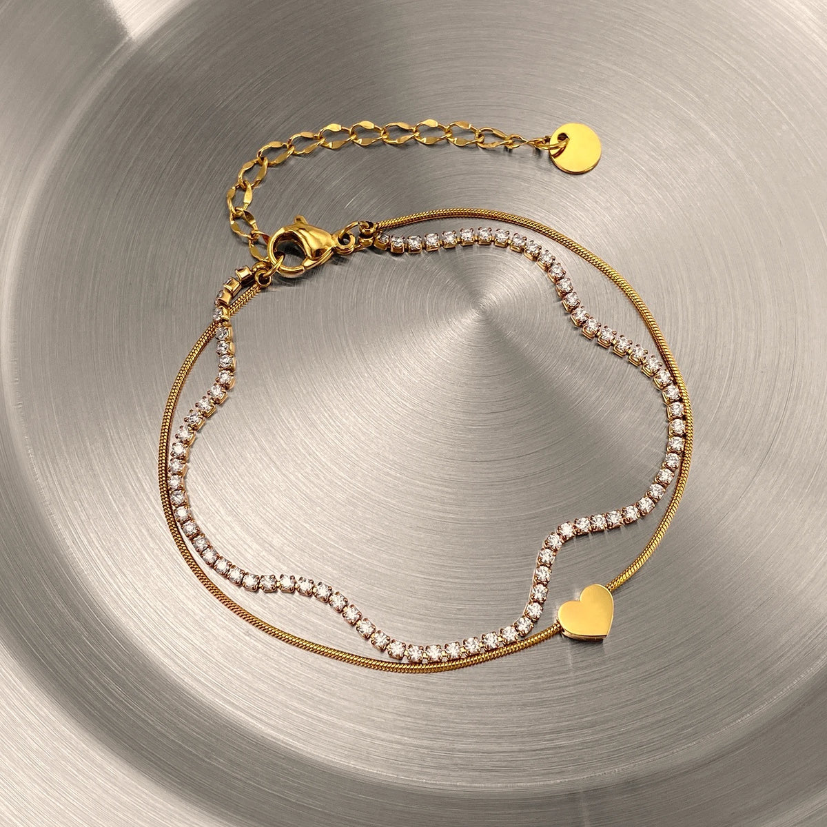 The Duo Gold Heart Charm Double Layer Bracelet — close-up flat lay on white linen showing heart charm, crystal tennis chain, and gold disc tag on extender