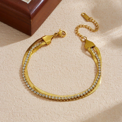 The Glide Gold Tennis & Herringbone Double Layer Bracelet — close-up flat lay on textured cream surface showing gold end cap detail and crystal tennis chain stones