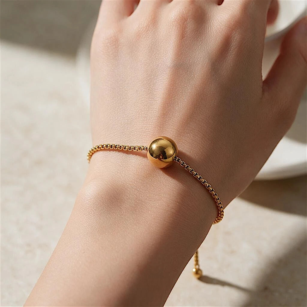 The Orbit Gold Sphere Slider Bracelet — styled flat on marble tray with dried white flowers showing full bracelet with slider closure detail and two ball-tipped tails