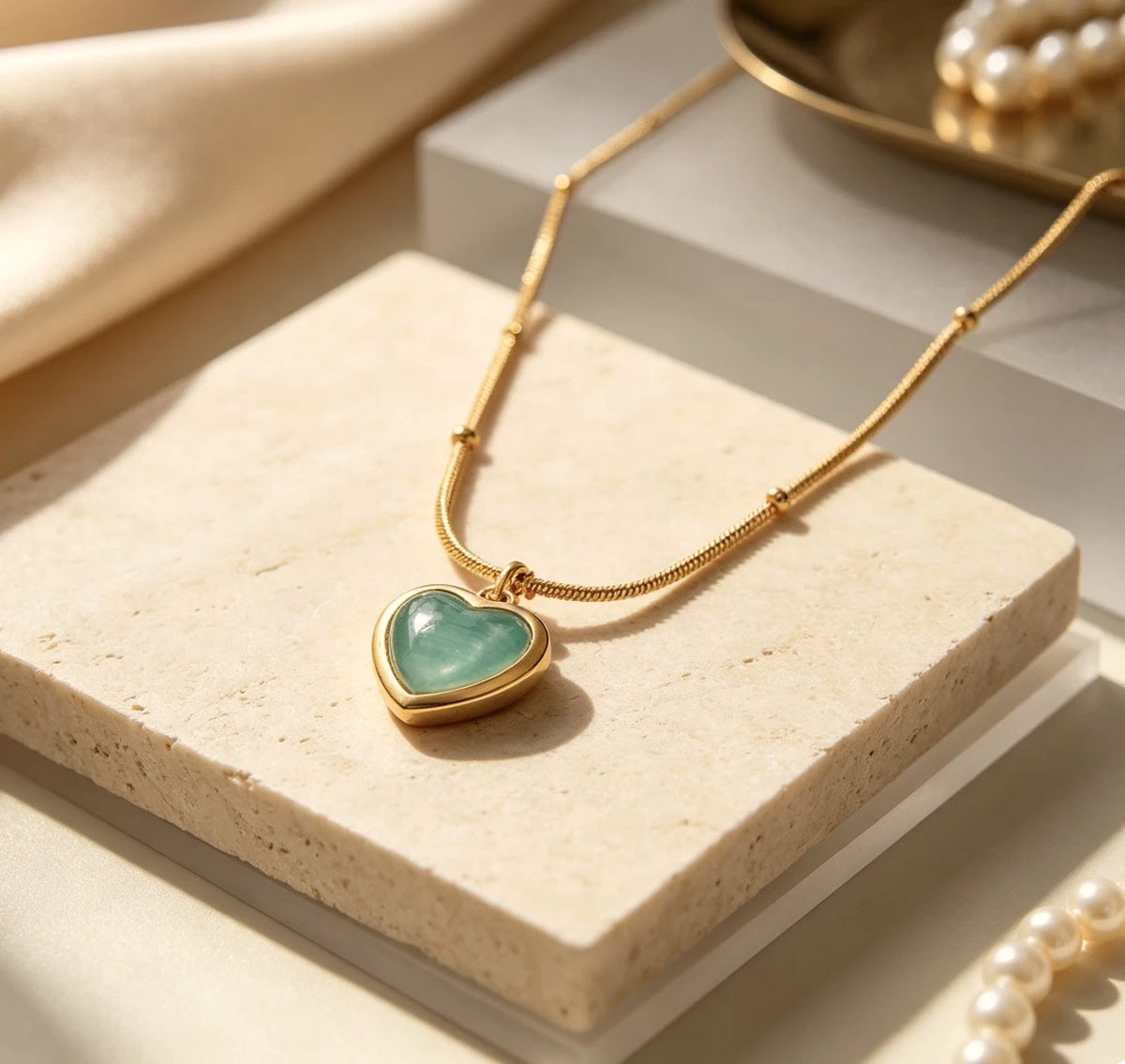 The Celeste Aqua Heart Cabochon Pendant Necklace on travertine marble with pearls – aqua heart pendant showing domed glassy cabochon surface in warm light