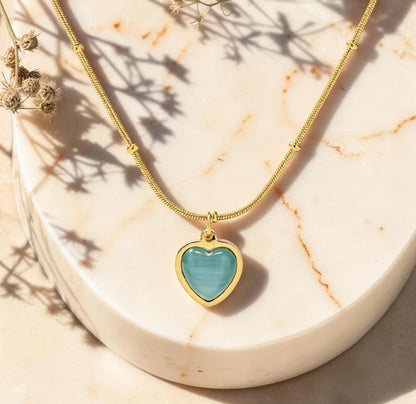The Celeste Aqua Heart Cabochon Pendant Necklace flat lay on marble with dried florals – bezel-set aqua heart pendant on gold satellite bead chain